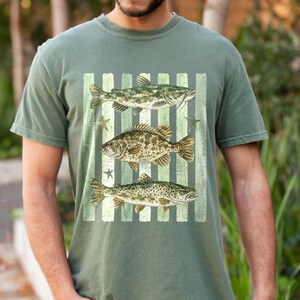 May include: Olive green t-shirt featuring a graphic of three fish: a catfish, a bass, and a trout, set against a striped background with star accents. The design is in shades of green and brown.