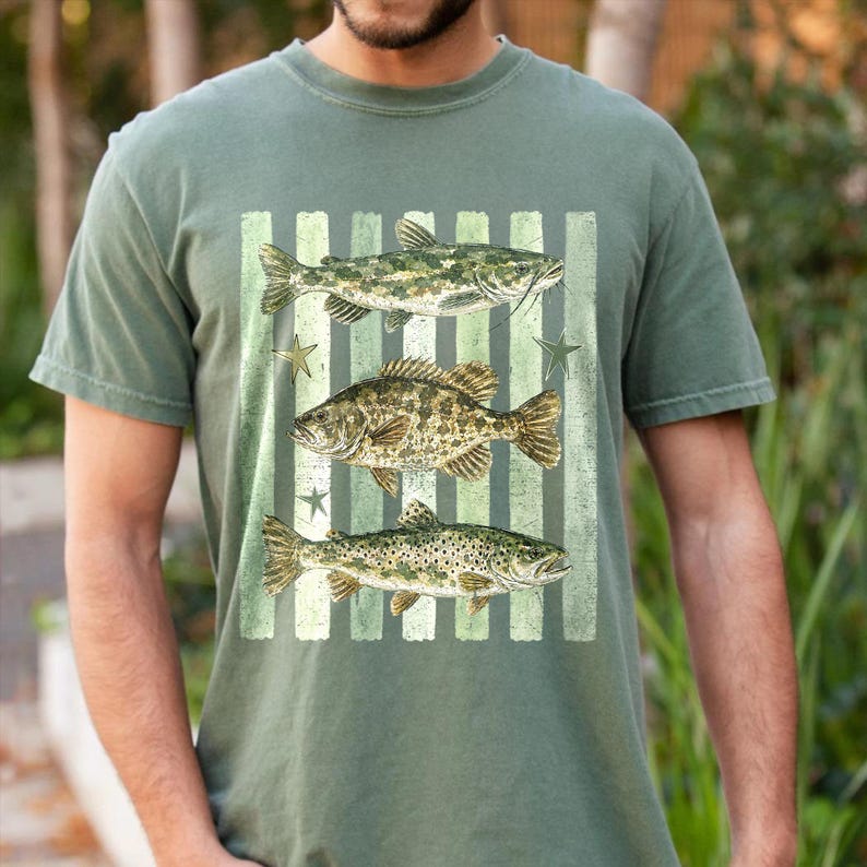 May include: Olive green t-shirt featuring a graphic of three fish: a catfish, a bass, and a trout, set against a striped background with star accents. The design is in shades of green and brown.