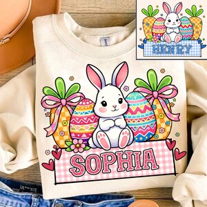 May include: Cream-colored sweatshirt featuring a cartoon Easter bunny with decorated eggs and the name "SOPHIA" in pink letters. The design includes carrots, flowers, and hearts, perfect for Easter celebrations.