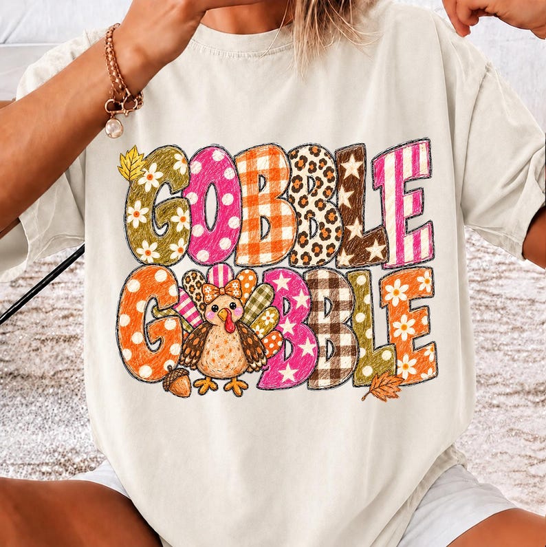 May include: A cream-colored short-sleeve t-shirt featuring the text GOBBLE GOBBLE in large, multi-patterned letters. The design includes a central cartoon turkey illustration, small floral motifs, leopard print, plaid, and star patterns.