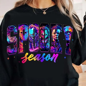 Puede incluir: Camiseta negra de manga larga con las palabras "Spooky Season" en una fuente colorida y estilizada. La palabra "Spooky" presenta imágenes de personajes de películas de terror. La palabra "season" está en un degradado arcoíris.