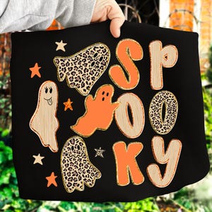 以下が含まれることがあります： 黒色のハロウィーンをテーマにしたデザインで、オレンジとタン色の文字で「SPOOKY」という言葉が書かれています。デザインには、ヒョウ柄模様のゴーストと星のグラフィックが含まれています。全体的なテーマは、お祭り騒ぎで不気味です。