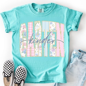 May include: A turquoise Comfort Colors t-shirt featuring the words "GARDEN" and "kinder" in a floral and patterned design. The shirt is styled with black and white checkered slip-on shoes and denim shorts.