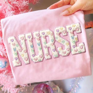May include: A pale pink t-shirt featuring the word "NURSE" in large, embroidered letters. The letters are filled with small, multi-coloured heart shapes. The embroidery has a textured, raised appearance.