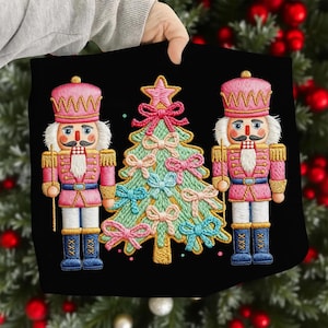 May include: Black embroidered cushion featuring two pink nutcrackers and a Christmas tree. The tree is decorated with pink and blue bows. The nutcrackers wear pink uniforms and blue boots, holding golden staffs.