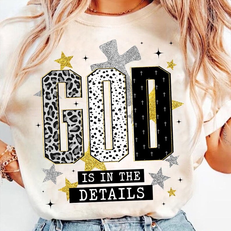 Op de afbeelding: Cr&egrave;mekleurig T-shirt met de tekst "GOD IS IN THE DETAILS" in een decoratief lettertype. De letters zijn in luipaardprint, stippen en zwart met gouden accenten. Zilveren en gouden sterren en kruisen maken het ontwerp compleet.