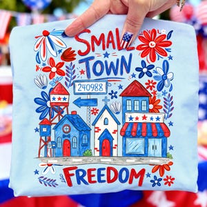 Może przedstawiać: Jasnoniebieska koszulka z kolorową grafiką przedstawiającą scenę z małego miasteczka. Projekt zawiera napisy "Small Town Freedom" oraz ulicę z budynkami, kwiatami i wieżą ciśnień. Grafika jest w kolorach czerwonym, białym i niebieskim.