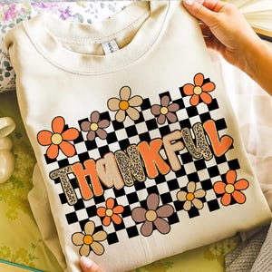 May include: Cream-colored sweatshirt with the word "THANKFUL" in orange and leopard print letters. The design includes a black and white checkered background and colorful flower graphics in shades of orange, brown, and tan.