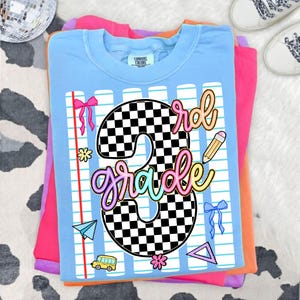 May include: A light blue t-shirt featuring a large black and white checkered number 3 with the word grade written in colorful script. The design includes a notebook paper background, a yellow school bus, a pencil, bows, and geometric shapes.