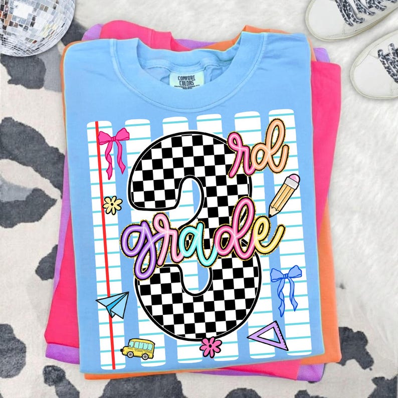 May include: A light blue t-shirt featuring a large black and white checkered number 3 with the word grade written in colorful script. The design includes a notebook paper background, a yellow school bus, a pencil, bows, and geometric shapes.