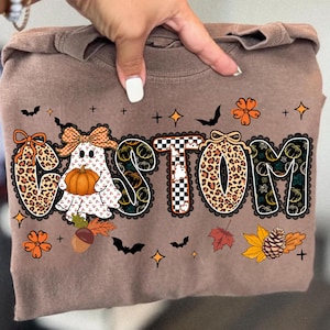 May include: A brown long-sleeve shirt with the word "CUSTOM" in a decorative font. The letters feature a ghost holding a pumpkin, leopard print, and pumpkin designs. The shirt is decorated with bats, stars, and fall foliage.