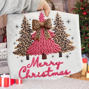 May include: White Christmas-themed pillow with embroidered pink Christmas tree, leopard print bow, and two leopard print trees. The words "Merry Christmas" are embroidered in pink. Black star accents complete the design.