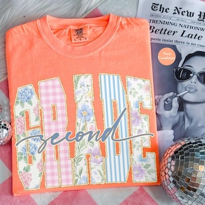 May include: A neon orange t-shirt with the words "GRADE Second TIME" in a decorative font. The letters are filled with floral and gingham patterns, outlined in gold. A newspaper and disco ball are in the background.