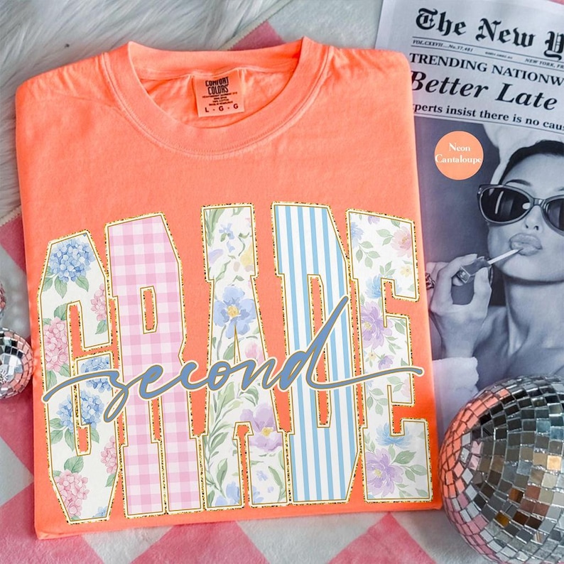 May include: A neon orange t-shirt with the words "GRADE Second TIME" in a decorative font. The letters are filled with floral and gingham patterns, outlined in gold. A newspaper and disco ball are in the background.