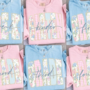 May include: Six crew neck sweatshirts in pastel shades of blue and pink. Each top displays a school grade level, like "Kinder" or "Third", in a floral and plaid pattern. A silver necklace is visible on each garment.