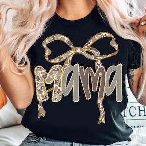 May include: Black t-shirt featuring the word "MAMA" in a layered design. The top layer of the letters has a camouflage pattern, and the bottom layer is a solid gray. A bow with a matching camouflage pattern sits above the word.