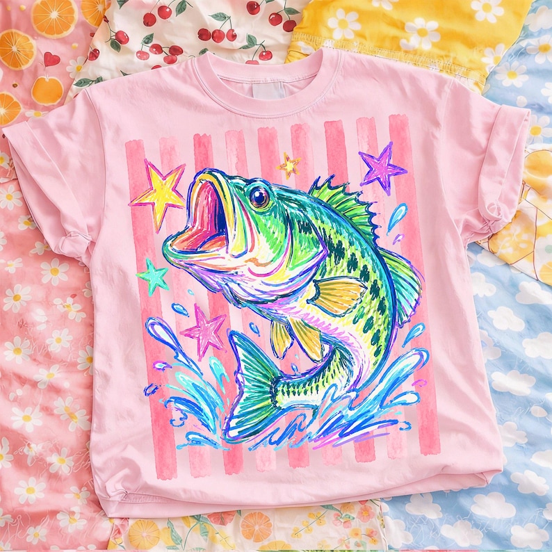 Op de afbeelding: Lichtroze t-shirt met een kleurrijke illustratie van een vis die uit het water springt. De vis is groen, blauw en geel, met een open mond. Roze verticale strepen en sterren staan achter de vis.