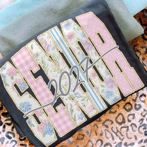 May include: Dark gray sweatshirt with the word "SENIOR" in a floral and polka dot design, with the year "2027" in the center. The sweatshirt has a gold outline and is folded. Other folded shirts are in the background.
