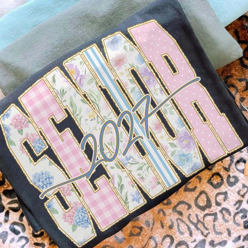 May include: Dark gray sweatshirt with the word "SENIOR" in a floral and polka dot design, with the year "2027" in the center. The sweatshirt has a gold outline and is folded. Other folded shirts are in the background.
