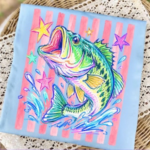 Op de afbeelding: Lichtblauw T-shirt met een kleurrijke illustratie van een baarsvis met open mond, omringd door sterren en waterspetters. Het ontwerp bevat roze verticale strepen. De vis is weergegeven in levendige groen-, blauw- en geeltinten.