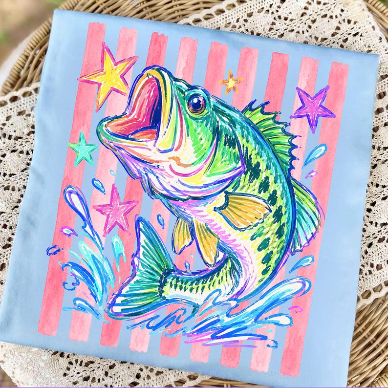 Op de afbeelding: Lichtblauw T-shirt met een kleurrijke illustratie van een baarsvis met open mond, omringd door sterren en waterspetters. Het ontwerp bevat roze verticale strepen. De vis is weergegeven in levendige groen-, blauw- en geeltinten.