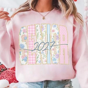 May include: A light pink sweatshirt with the word "SENIOR" in large, gold-outlined letters. The letters are filled with floral, gingham, and polka dot patterns. The year "2027" is written in a cursive font across the center of the word.
