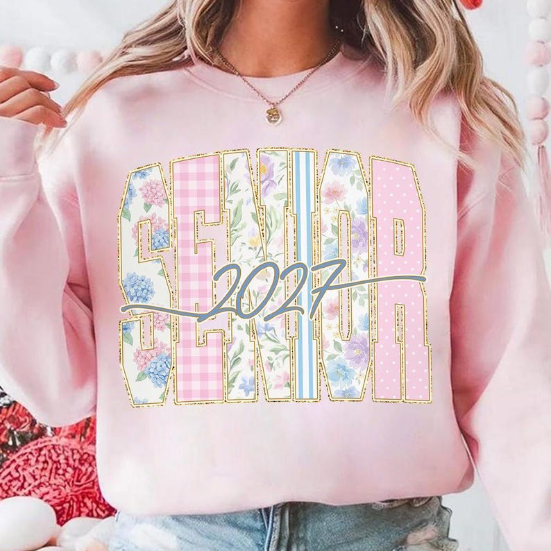 May include: A light pink sweatshirt with the word "SENIOR" in large, gold-outlined letters. The letters are filled with floral, gingham, and polka dot patterns. The year "2027" is written in a cursive font across the center of the word.