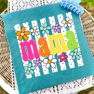 May include: Teal pillow with the word "MAMA" in colorful bubble letters, surrounded by flowers and white stripes with black polka dots. A blue hair clip is in the upper right corner.
