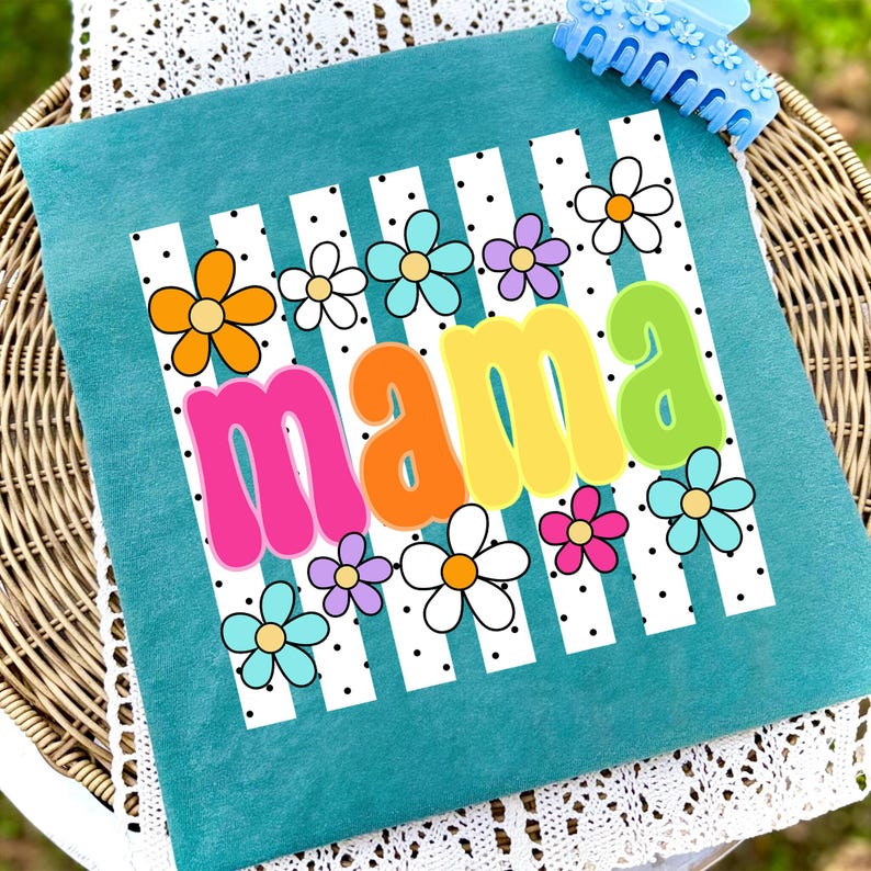 May include: Teal pillow with the word "MAMA" in colorful bubble letters, surrounded by flowers and white stripes with black polka dots. A blue hair clip is in the upper right corner.