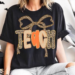 May include: Black t-shirt featuring the word "TEACH" in a unique design. The letters are outlined in a light brown color with an orange center. A leopard print bow is above the word, and the letters are also leopard print.