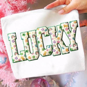 May include: White square fabric with the word "LUCKY" embroidered in large, textured letters. The letters are filled with colorful, whimsical designs and outlined in green. The fabric is held by a hand, with a pink and white background.