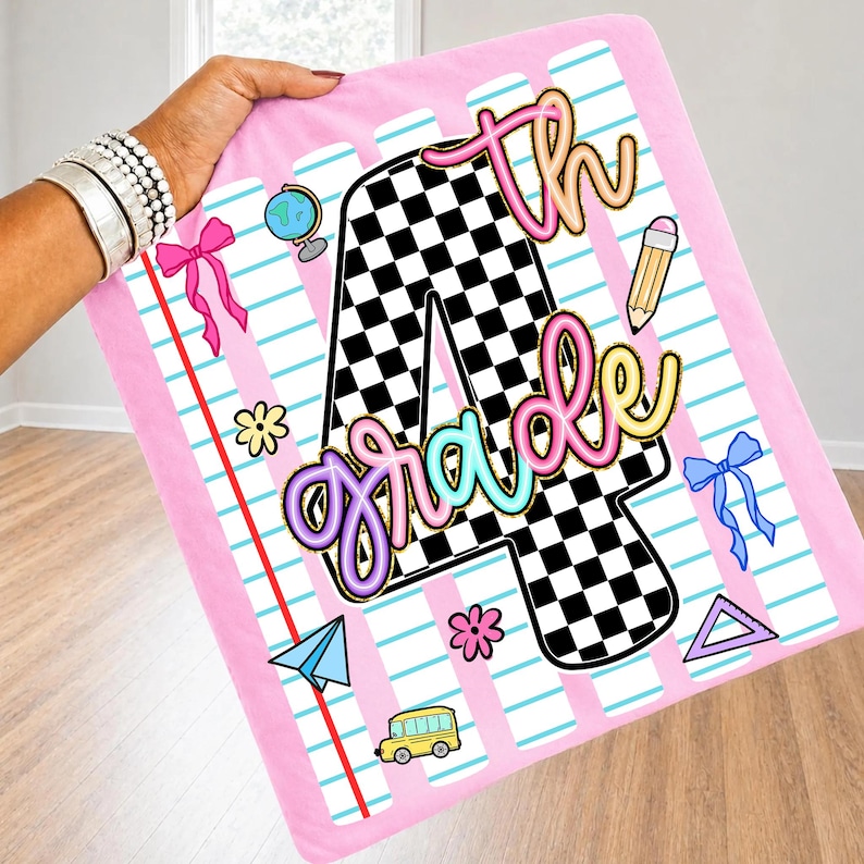 May include: A pink blanket featuring a large black and white checkered number 4 with the text 4th grade in neon-colored script. The design includes school-themed graphics like a globe, pencil, paper airplane, school bus, and bows on lined paper.