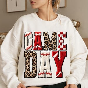 May include: White sweatshirt with the words "GAME DAY" in large, bold letters. The letters have baseball stitching and leopard print accents. A fun and sporty design for game day.