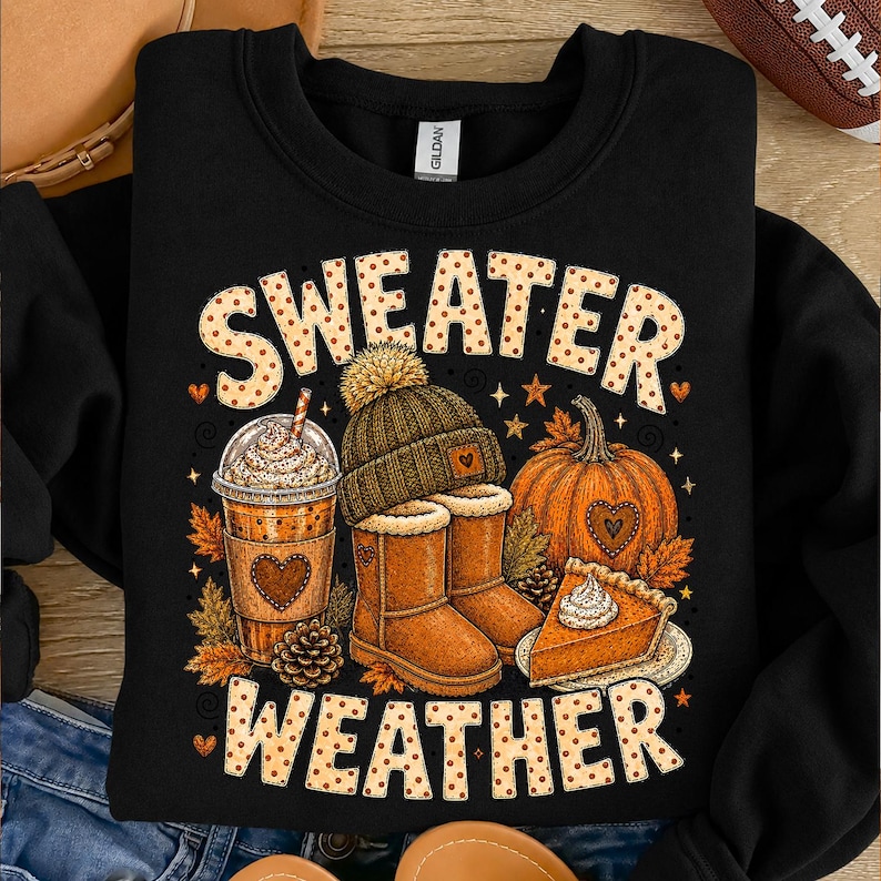 May include: A black crewneck sweatshirt featuring a graphic print with the text "SWEATER WEATHER" in a tan polka-dot font. The design includes a coffee cup, a knit beanie, a pair of tan boots, a pumpkin, and a slice of pumpkin pie with whipped cream.