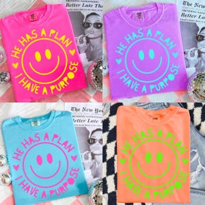 Puede incluir: Cuatro camisetas coloridas en rosa, morado, azul y naranja, cada una con un dise&ntilde;o de carita sonriente y el texto "HE HAS A PLAN, I HAVE A PURPOSE". Las camisetas se muestran con bolas de discoteca.