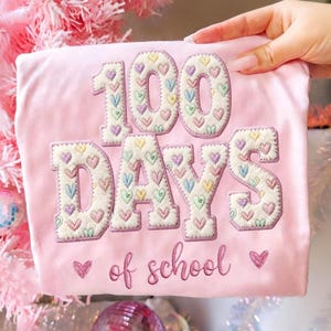 Puede incluir: Una camiseta rosa claro con la frase bordada "100 DAYS of school". Los números y letras están rellenos de corazones coloridos. Dos pequeños diseños de corazón están debajo del texto.