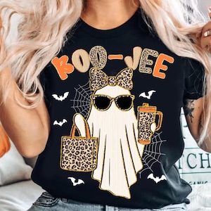 May include: Black t-shirt with a Halloween ghost graphic. The ghost wears sunglasses and a bow, carrying a tote bag and a mug. The text "BOO-JEE" is in orange and leopard print.