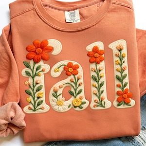 May include: Coral-colored sweatshirt with the word "Fall" embroidered in cream-colored, floral-decorated letters. The flowers are orange, yellow, and cream, with green stems and leaves. The sweatshirt has a crew neck and long sleeves.