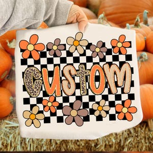 May include: A white tote bag featuring the word "CUSTOM" in orange and leopard print lettering, surrounded by orange, brown, and tan flowers. A black and white checkered pattern fills the background.