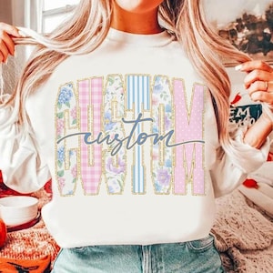 May include: White long-sleeve sweatshirt with the word "CUSTOM" in a colorful, floral, and patterned design. The word "custom" is written in a cursive font across the center of the word "CUSTOM".