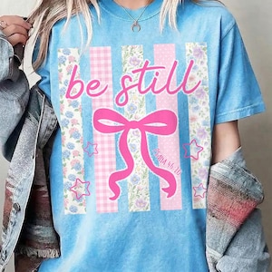 K&ouml;nnte beinhalten: Hellblaues T-Shirt mit dem rosa Schriftzug "be still". Das Design umfasst eine rosa Schleife, Sterne und vertikale Streifen mit Blumen- und Vichy-Mustern. Der Text "Psalm 46:10" ist ebenfalls vorhanden.