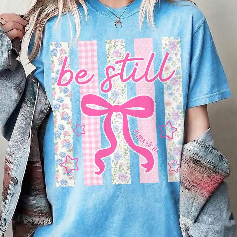 K&ouml;nnte beinhalten: Hellblaues T-Shirt mit dem rosa Schriftzug "be still". Das Design umfasst eine rosa Schleife, Sterne und vertikale Streifen mit Blumen- und Vichy-Mustern. Der Text "Psalm 46:10" ist ebenfalls vorhanden.