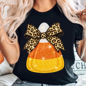 May include: Black t-shirt featuring a candy corn graphic with a leopard print bow. The candy corn design has orange, yellow, and white sections with glitter accents. The bow has a gold glitter outline.