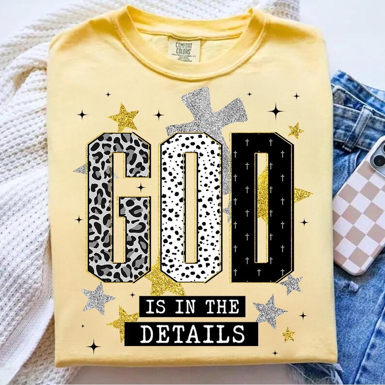 Op de afbeelding: Geel Comfort Colors T-shirt met de tekst "GOD IS IN THE DETAILS". Het woord "GOD" heeft een luipaardprint, stippen en een zwart ontwerp. Zilveren en gouden sterren en een zilveren kruis zijn ook te zien.