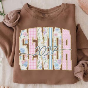 May include: A brown sweatshirt with the word "SENIOR" in a floral and patterned design, with the year "2027" in the center. The letters are outlined in gold. The design includes pink gingham, blue stripes, and floral patterns.