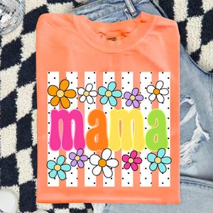 May include: An orange crew neck t-shirt featuring a graphic print of the word mama in colorful bubble letters. The design includes a background of vertical white stripes with black polka dots and various multi-colored flower illustrations.