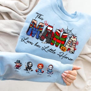 Pu&ograve; includere: Felpa azzurra con la scritta "This Mama Loves her Little Horror". La parola "Mama" &egrave; disegnata con personaggi di film horror. La manica presenta quattro piccole illustrazioni di personaggi horror con nomi.