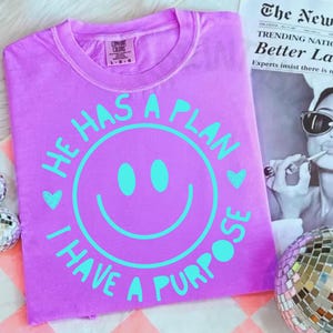 Puede incluir: Una camiseta lila con un dise&ntilde;o de cara sonriente turquesa. El texto "HE HAS A PLAN" y "I HAVE A PURPOSE" est&aacute;n en turquesa alrededor de la cara sonriente. La camiseta est&aacute; doblada y tiene una etiqueta Comfort Colors.