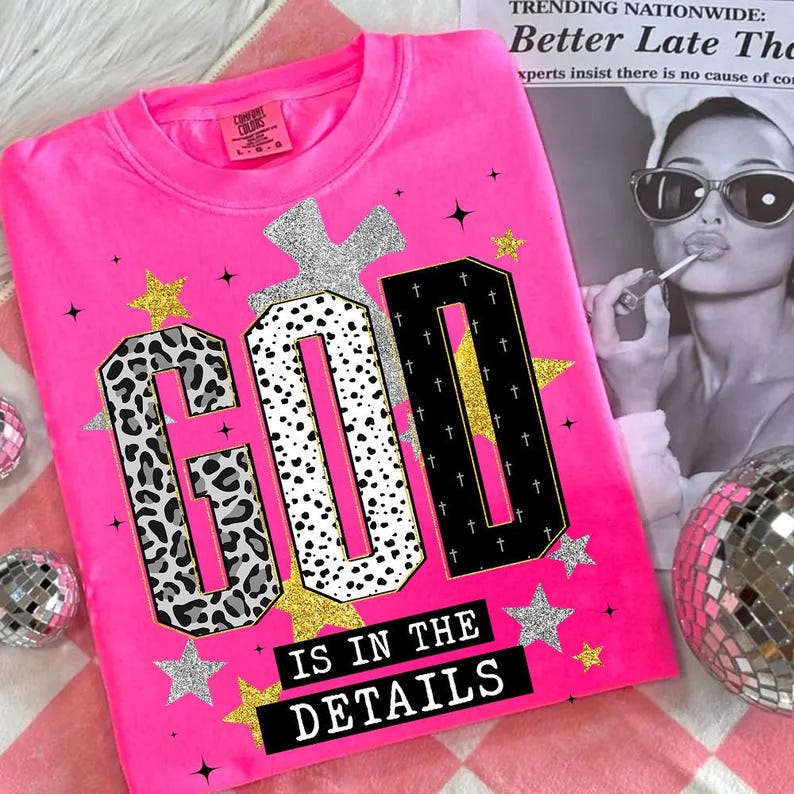 Op de afbeelding: Een felroze sweatshirt met de tekst "GOD IS IN THE DETAILS" in een vet, decoratief lettertype. De letters hebben luipaardprint, stippen en kruisontwerpen. Gouden en zilveren sterren en een zilveren kruis accentueren het ontwerp.