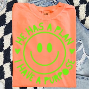 Puede incluir: Una camiseta de color coral con un dise&ntilde;o de carita sonriente verde ne&oacute;n. El texto alrededor de la carita sonriente dice "HE HAS A PLAN, I HAVE A PURPOSE". La camiseta est&aacute; doblada y colocada sobre unos vaqueros azules.